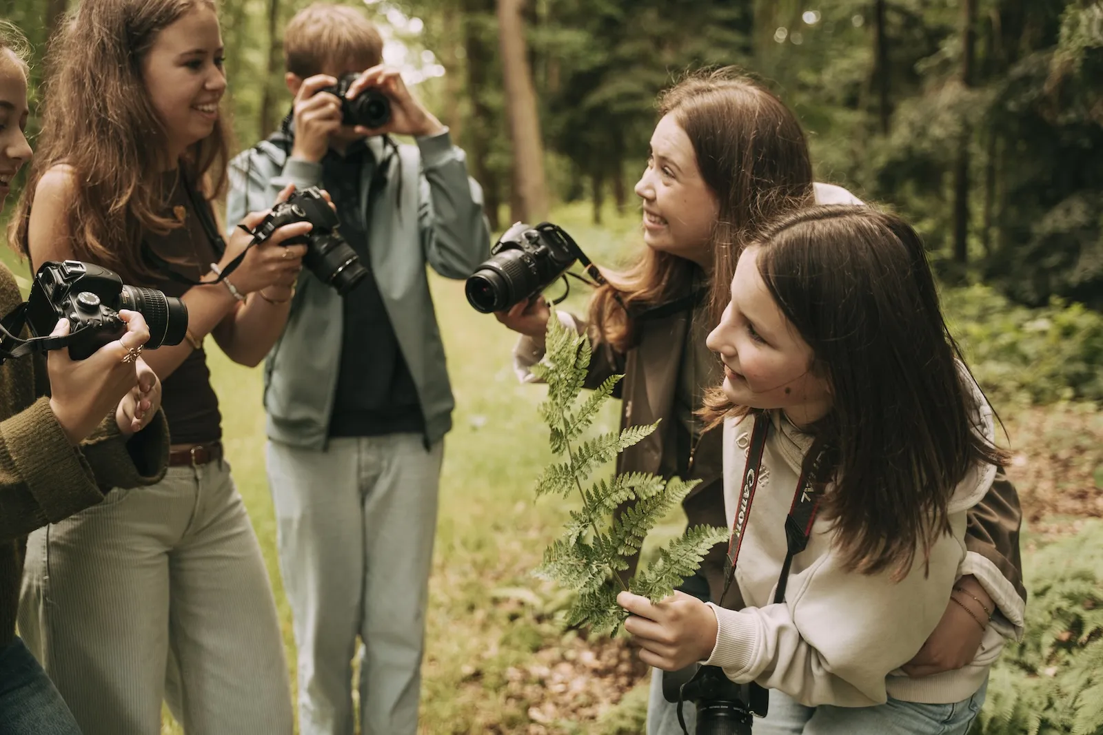 Kinderen en een begeleidster bekijken samen een varenblad in het bos, met spiegelreflex- en systeemcamera's in de hand