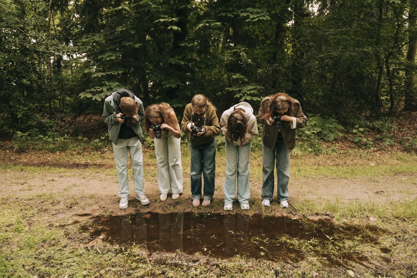 Vijf kinderen staan op een rij voorovergebogen boven een plas en fotograferen de weerspiegeling in het water