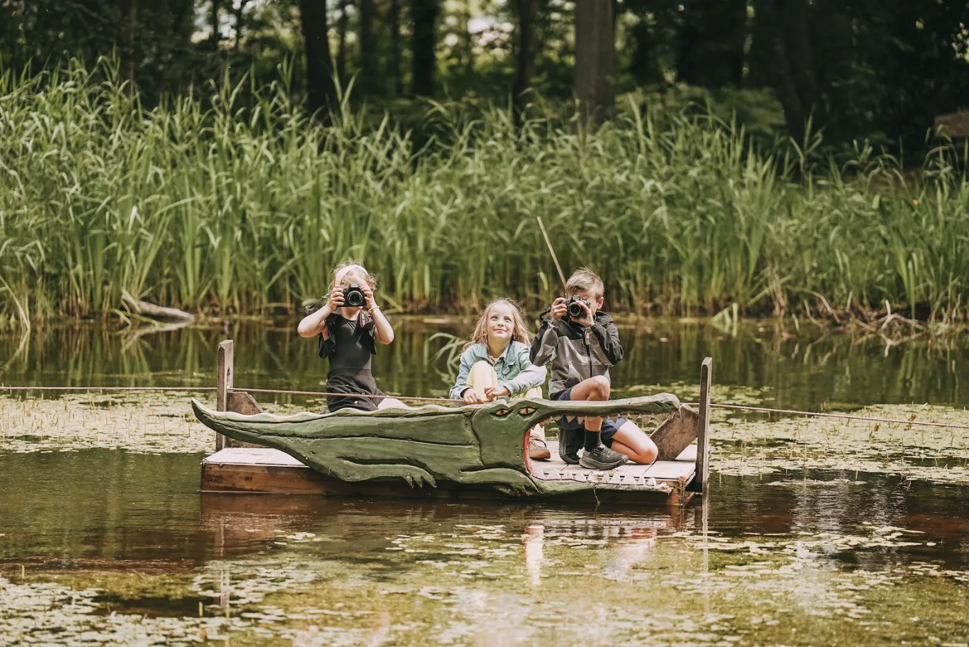Drie kinderen op een zelfgebouwd trekvlot in de vorm van een krokodil midden op een vijver; twee fotograferen omhoog met hun camera
